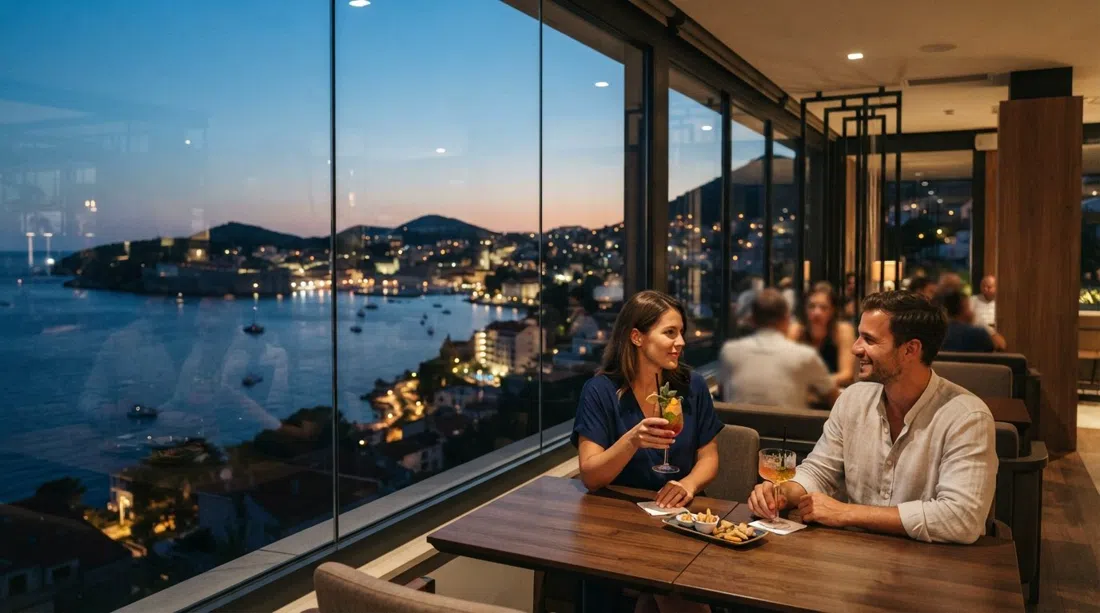 el Zenith Bar en la última planta del Hotel Kompas al anochecer, ventanales de suelo a techo con vistas a la bahía de Lapad, cócteles y luces de la ciudad al fondo
