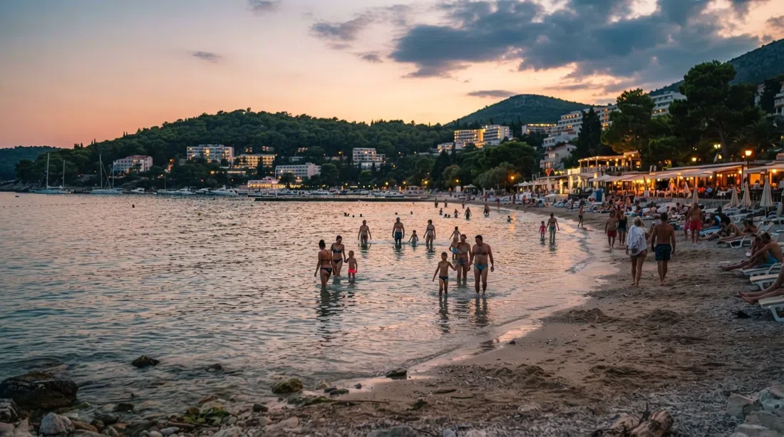 Uvala Lapad al atardecer, aguas tranquilas y poco profundas, familias vadeando lejos de la orilla y bares de playa iluminados a lo largo de la media luna de guijarros y arena